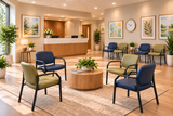 A bright, modern reception lobby with multiple upholstered chairs in blue and olive green arranged around a round wooden coffee table, with a reception desk, framed wall art, and potted plants in the background.